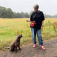 A woman standing on a dirt path in an open grassy field, wearing a black oversized T-shirt, blue jeans, and red sandals, with a small red and black crossbody bag. A brown curly-haired dog is sitting beside her, looking up.