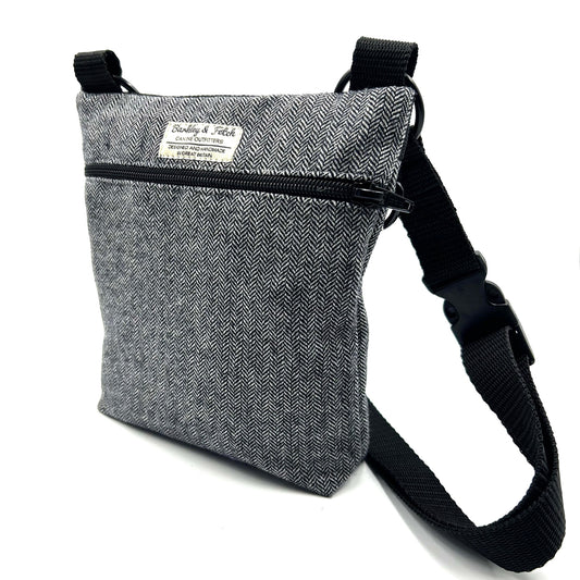 A gray herringbone patterned dog walk and treat bag with an adjustable black shoulder strap and a zip fastening.