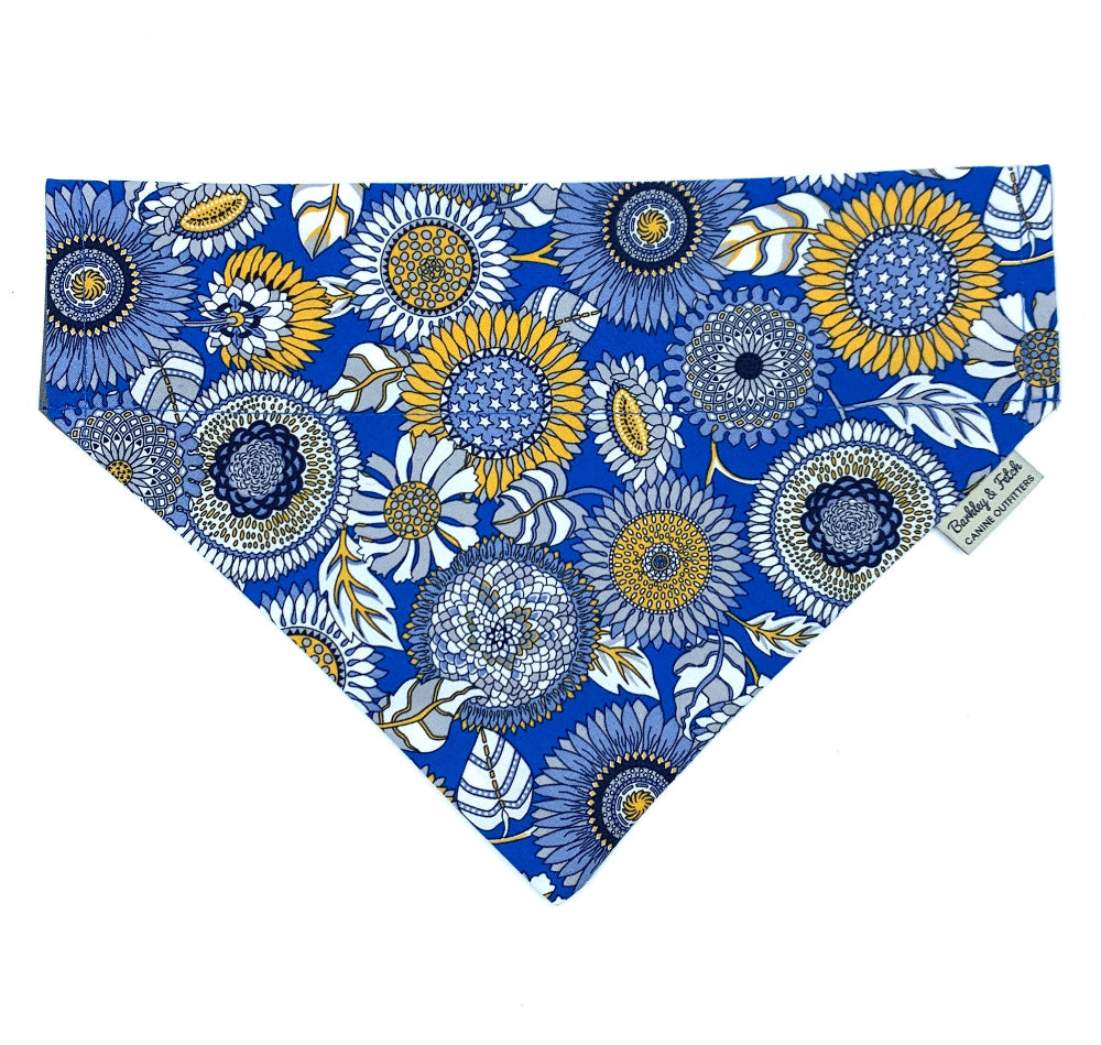 A blue dog bandana with a floral pattern, including large sunflower-like designs and smaller floral accents in yellow and white.