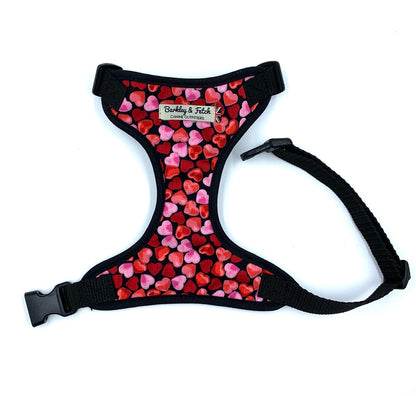 A dog harness with a love hearts print, predominantly red and pink, with adjustable straps and a metal d-ring for leash attachment.